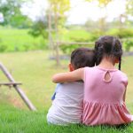 Older sister hugs little brother by the neck, shoulders sitting on green grass field. Two adorable Asian children sitting and hugging the neck rear view.