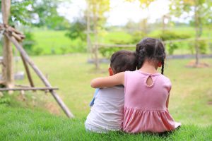 Older sister hugs little brother by the neck, shoulders sitting on green grass field. Two adorable Asian children sitting and hugging the neck rear view.